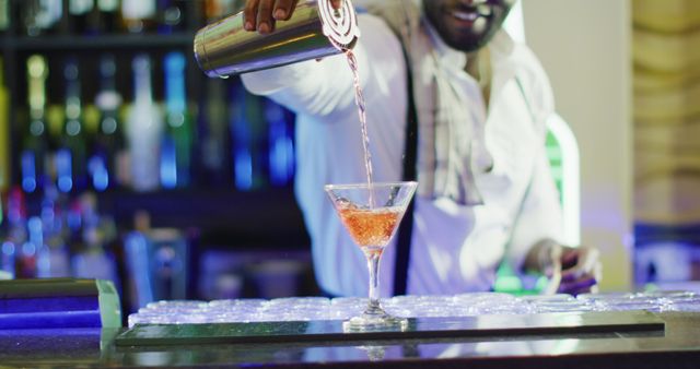 Bartender pouring crafted cocktail into martini glass at a bar counter, suitable for illustrating themes such as nightlife, hospitality, and professional bartending. Ideal for use in advertisements, marketing materials for bars, clubs, or restaurants, and content related to mixology or beverage services.