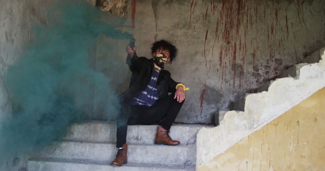 Person sitting on staircase in dilapidated building with graffiti holding smoke bomb, creating dense blue-green smoke. Suitable for themes like mystery, urban exploration, street style, rebellion, desolation, and artistic photography.