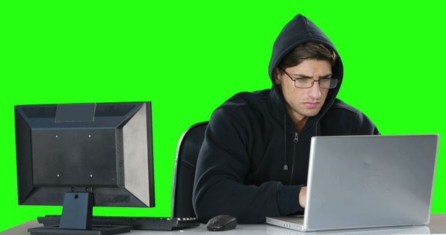 A young Caucasian man in a hoodie is focused on his laptop, with copy space on the green background. His intense concentration suggests he could be a programmer or gamer engaged in his work or play.