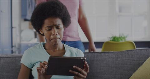 Teen tapping tablet on sofa while parent watching nearby in sunlit living room