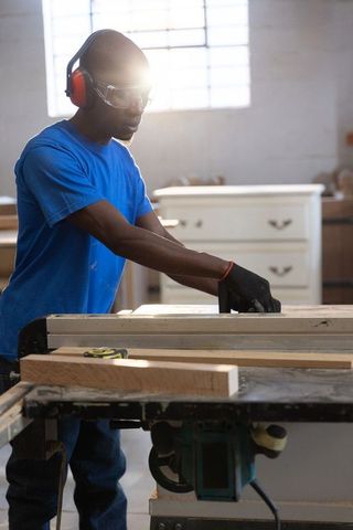 Skilled woodworker cutting lumber on table saw with safety gear, sunlit carpentry workshop