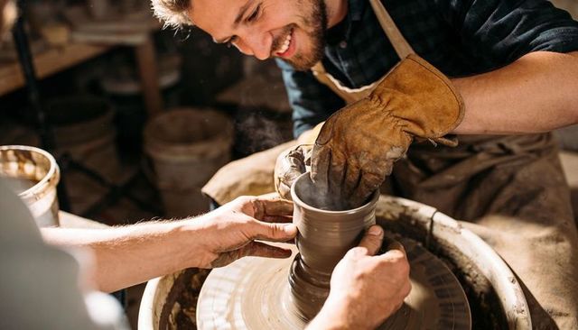 Smiling potter mentoring student while shaping clay on pottery wheel in rustic studio