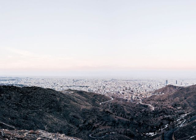 This panoramic cityscape captures the urban expanse from a high mountain viewpoint at dusk. The natural landscape contrasts with the dense urban skyline, providing a sense of calm and serenity. Suitable for use in travel brochures, city guides, urban development presentations, or as a scenic desktop wallpaper.
