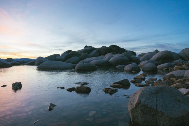 Rocky shoreline with large boulders scattered around and reflected sky in calm water at sunset. Serene atmosphere with a gradient blue sky transitioning to sunset colors. Useful for nature themes, travel blogs, background images for presentations, or screen savers emphasizing tranquility and the beauty of natural landscapes.