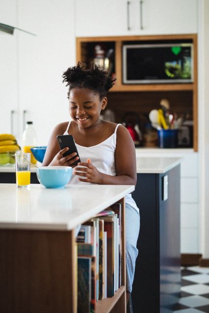 Young African American girl enjoying breakfast at home while using a smartphone. Ideal for themes related to childhood, healthy eating, modern lifestyle, and family life. Suitable for use in advertisements, blogs, and articles focusing on technology use among children, morning routines, and home life.