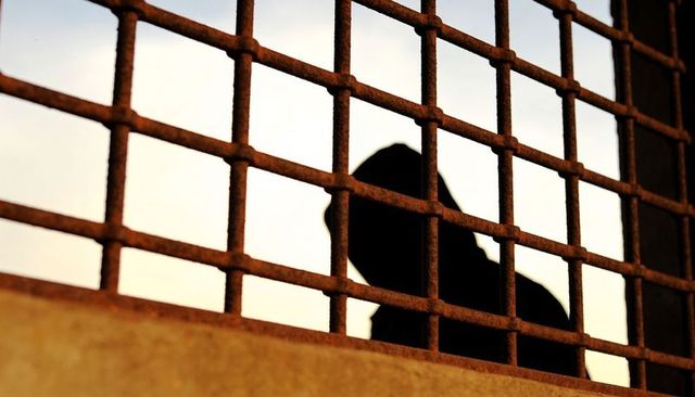Silhouetted hooded figure behind rusted grille on urban bridge, moody backlit isolation