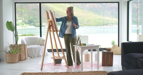 Mature woman painting at easel in sunlit waterside living room wearing headphones