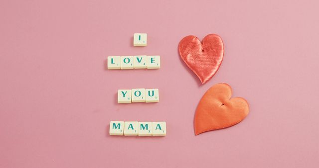 Scrabble tiles spell out I LOVE YOU MAMA on a pink background, flanked by two fabric hearts, with copy space. It's a warm, affectionate gesture, often associated with Mother's Day or expressing gratitude and love to mothers.