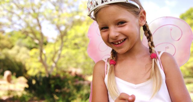 A young Caucasian girl dressed in a fairy costume with wings and a tiara smiles brightly in a sunny outdoor setting, with copy space. Her playful attire and joyful expression capture the innocence and imagination of childhood.