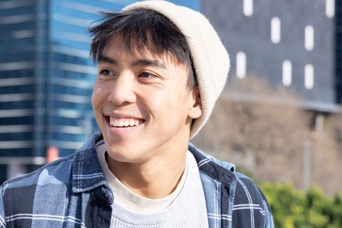 Sunlit smiling asian man wearing beanie and plaid shirt on urban street near glass office