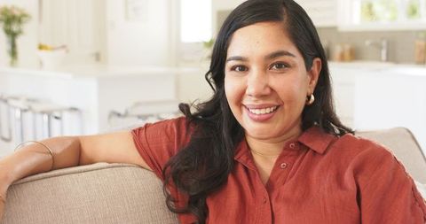 Smiling woman relaxing on beige sofa in bright contemporary kitchen wearing rust shirt
