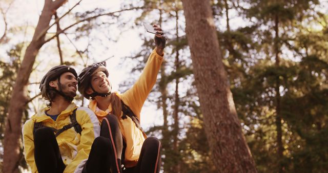 A young Caucasian couple wearing helmets and yellow jackets are taking a selfie during a bike ride in a forest, with copy space. Their joyful expressions and the outdoor setting suggest an adventurous and active lifestyle.