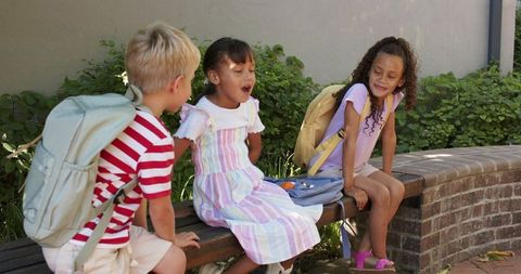 Diverse children sitting on bench with backpacks in sunny school courtyard, friends trio