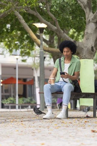 African american man sitting on bench holding coffee and checking smartphone in urban plaza