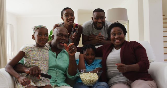 Multi-generational African American family gathered in the living room watching a movie together, displaying joyful and excited expressions. They have a bowl of popcorn and are sitting comfortably on the couch. Perfect for advertisements, blog posts, or articles focusing on family bonding, entertainment, and cultural representation.