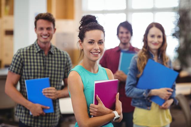 Young professionals standing in a modern office holding notebooks, smiling and looking confident. Ideal for use in business, teamwork, and collaboration contexts, as well as for promoting modern work environments and professional development.