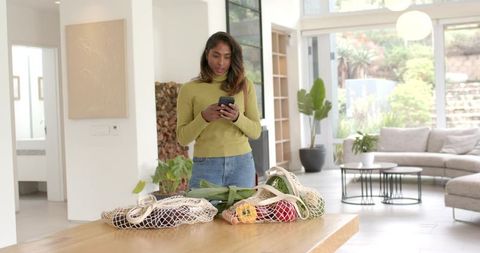 Indian woman holding phone checking reusable mesh grocery bags on modern kitchen island
