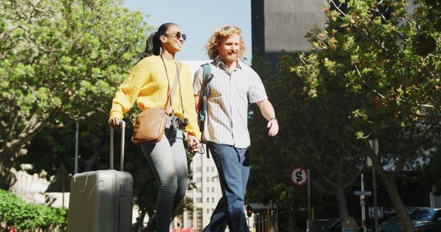 Happy diverse couple walking with luggage in sunny city street. City break, travel and hospitality concept.