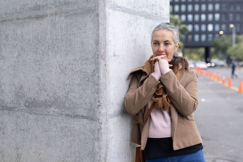 Contemplative mature woman leaning on concrete pillar wearing tan coat and scarf