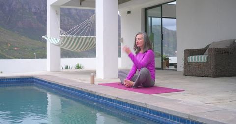 Mature woman practicing meditation poolside on magenta mat with mountain view
