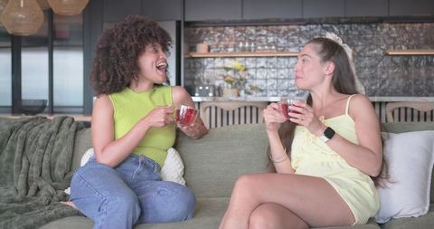 Two women laughing on green sofa sipping herbal tea in modern open-plan living room