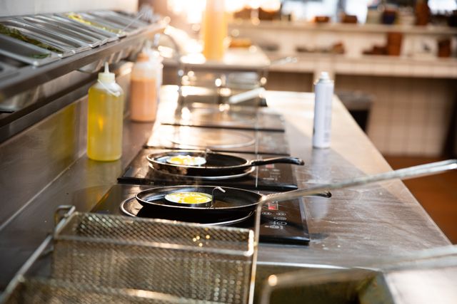 This image shows two frying pans on a stove with eggs frying in a professional kitchen. A bottle of olive oil is nearby. Ideal for use in culinary blogs, restaurant websites, cooking tutorials, and food-related advertisements.