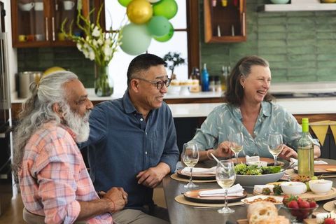 Diverse seniors sharing wine and salad at home dinner party, laughing around table