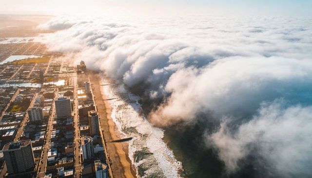 Dense fog bank rolling over beachfront skyline at sunrise, aerial coastal seascape