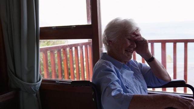 Elderly woman sits in wheelchair with a hand on forehead. Appears emotional with thoughtful expression near window showcasing landscape view outside. Use this to convey themes of aging, emotion, or senior isolation in blogs, healthcare publications, or social awareness campaigns.