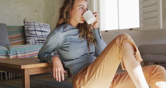 Thoughtful caucasian woman sitting on floor in living room drinking coffee, looking out of window. simple living in an off the grid rural home.