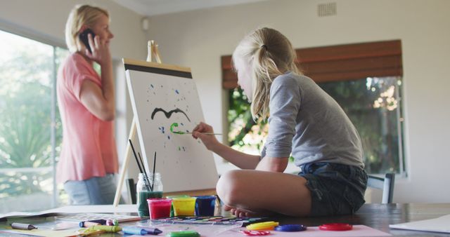 This scene shows a young girl sitting on a table and painting on an easel, with various colors of paint in front of her. In the background, a woman stands talking on the phone. The child is focused on her artwork, while the woman multitasks between family and personal responsibilities. Ideal use cases include topics on family life, home activities, mother-daughter bonding, and balancing responsibilities.