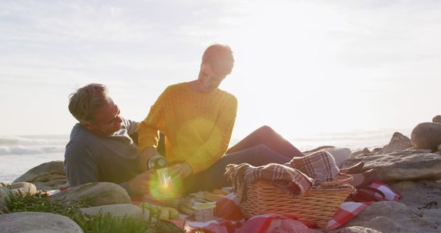 Couple is enjoying a peaceful picnic on a rocky beach during sunset. They seem relaxed and content, with a picnic basket and blanket spread out. Ideal for illustrating concepts of outdoor leisure, romantic getaways, nature appreciation, and summer activities.