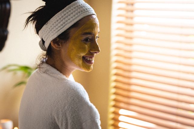 Smiling biracial young woman with headband and face pack looking away in spa. unaltered, vitiligo, spa, body care, skin care, bathroom, lifestyle and wellness.