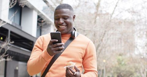 African American man smiling while using smartphone and holding muffin on city sidewalk