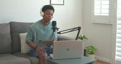 African american man recording podcast at home using laptop, microphone, headphones and tablet