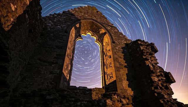 Gothic ruin window framing star trails circling night sky over crumbling stone tracery