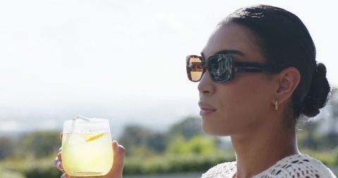 African woman wearing crochet top sipping lemon drink on sunny terrace with sunglasses