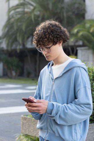 Teenager checking smartphone on sidewalk wearing light blue hoodie and glasses