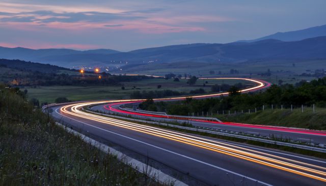 View featuring a winding highway with light trails from passing vehicles set against a rural landscape at dusk. Suitable for themes related to transportation, travel, adventure, and scenic views. Ideal for use in brochures, travel guides, websites, and marketing materials emphasizing road trips and countryside beauty.