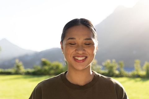 Sunlit asian woman smiling with eyes closed on mountain lawn, freckled, serene portrait