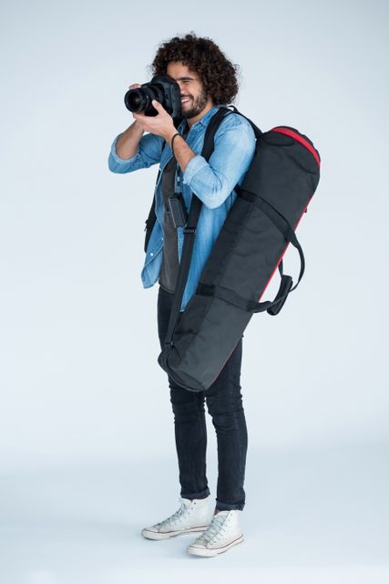Young male photographer with digital camera and tripod bag on his back, posing in studio. Ideal for content about photography, professional photographers, studio setup, and photography accessories.