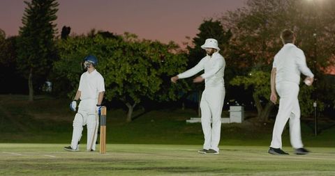 Twilight cricket scene with batsman at crease, bowler preparing, fielder standing