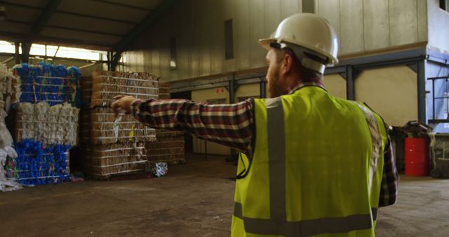 Warehouse manager in safety vest and hard hat pointing while inspecting stacked cardboard and plastic bales in an industrial facility. Useful for representations of logistics, recycling, industrial processes, warehouse management, and quality control.