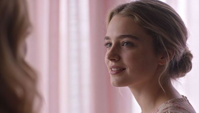 Young woman smiling in low bun leaning toward friend by pastel pink curtains soft daylight