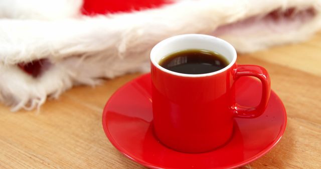 A red cup of coffee sits on a wooden table, with a hint of festive Santa Claus attire in the background, with copy space. The vibrant color of the cup adds a warm touch to the holiday atmosphere.