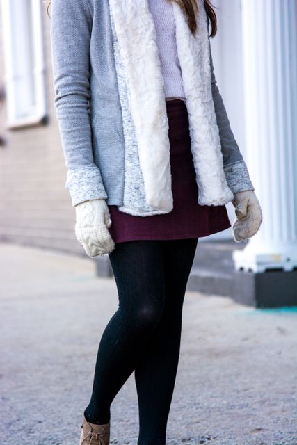 Young woman dressed in a stylish winter outfit, featuring a grey sweater with a furry lining, a maroon skirt, black tights, and white mittens. Suitable for use in fashion promotions, winter clothing advertisements, or lifestyle blogs focused on seasonal fashion trends.