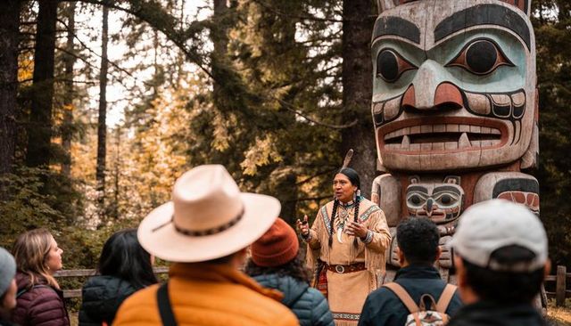 Storyteller in traditional regalia speaking to visitors by carved totem pole in autumn forest