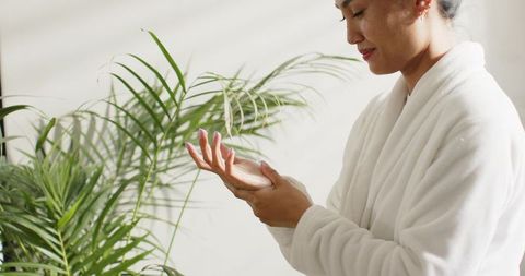 Mid adult asian woman applying moisturizer in white bathrobe beside sunlit palm plant