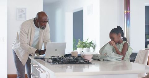 Senior man helping child pack backpack while working remotely at bright kitchen island