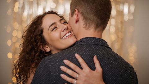 Smiling couple embracing under warm bokeh lights revealing engagement ring